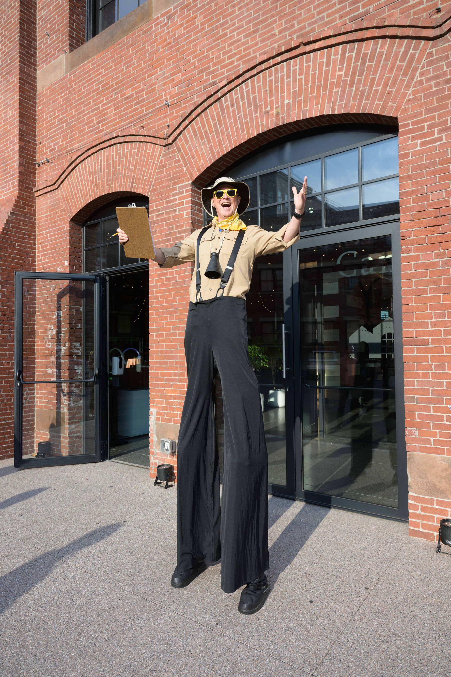 Person on stilts in front of a brick building, wearing a hat, suspenders, and waving with arms outstretched.