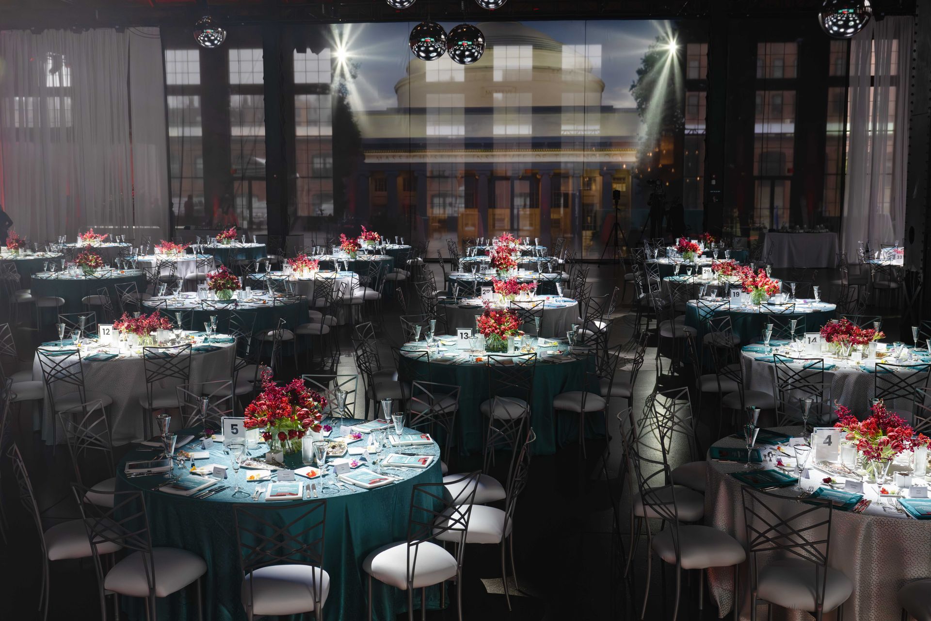 Elegant ballroom set for an event, round tables with teal and white linens, red floral centerpieces, black chairs, and light beams.