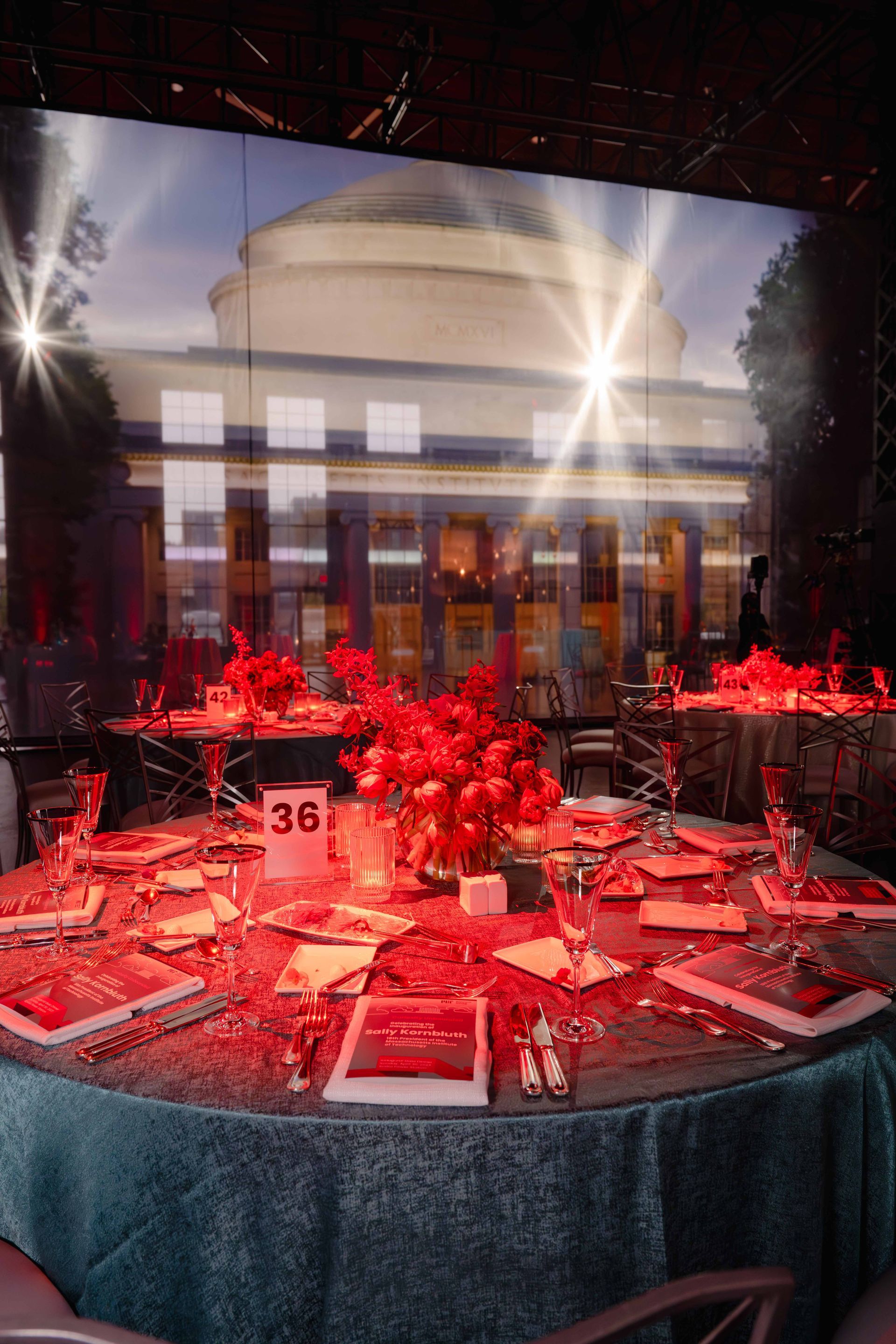 Formal dining table with red flowers, setting overlooking a building at dusk with streaks of light.