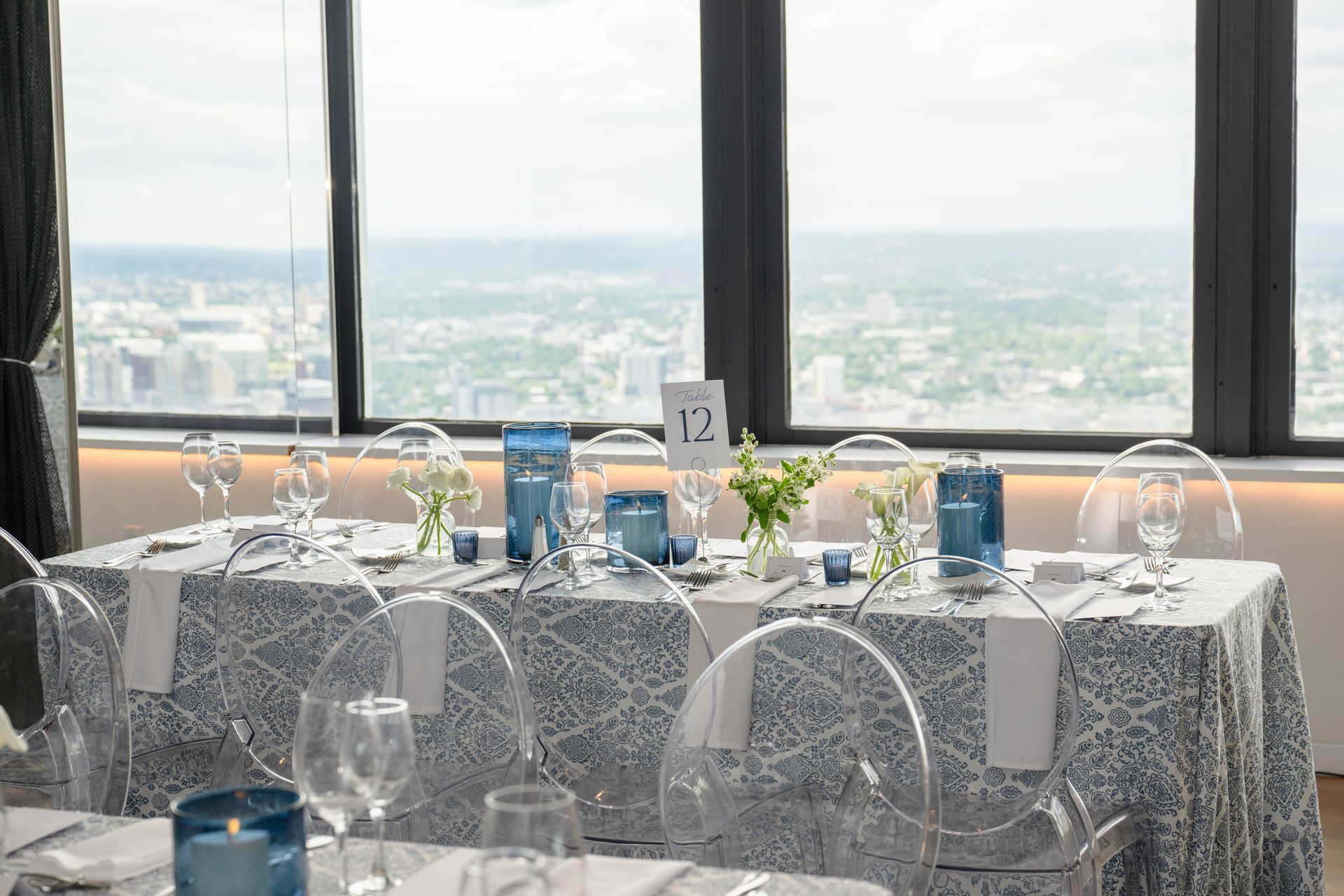 Elegant table setting with blue accents, a cityscape backdrop, and clear chairs. Table number 12.
