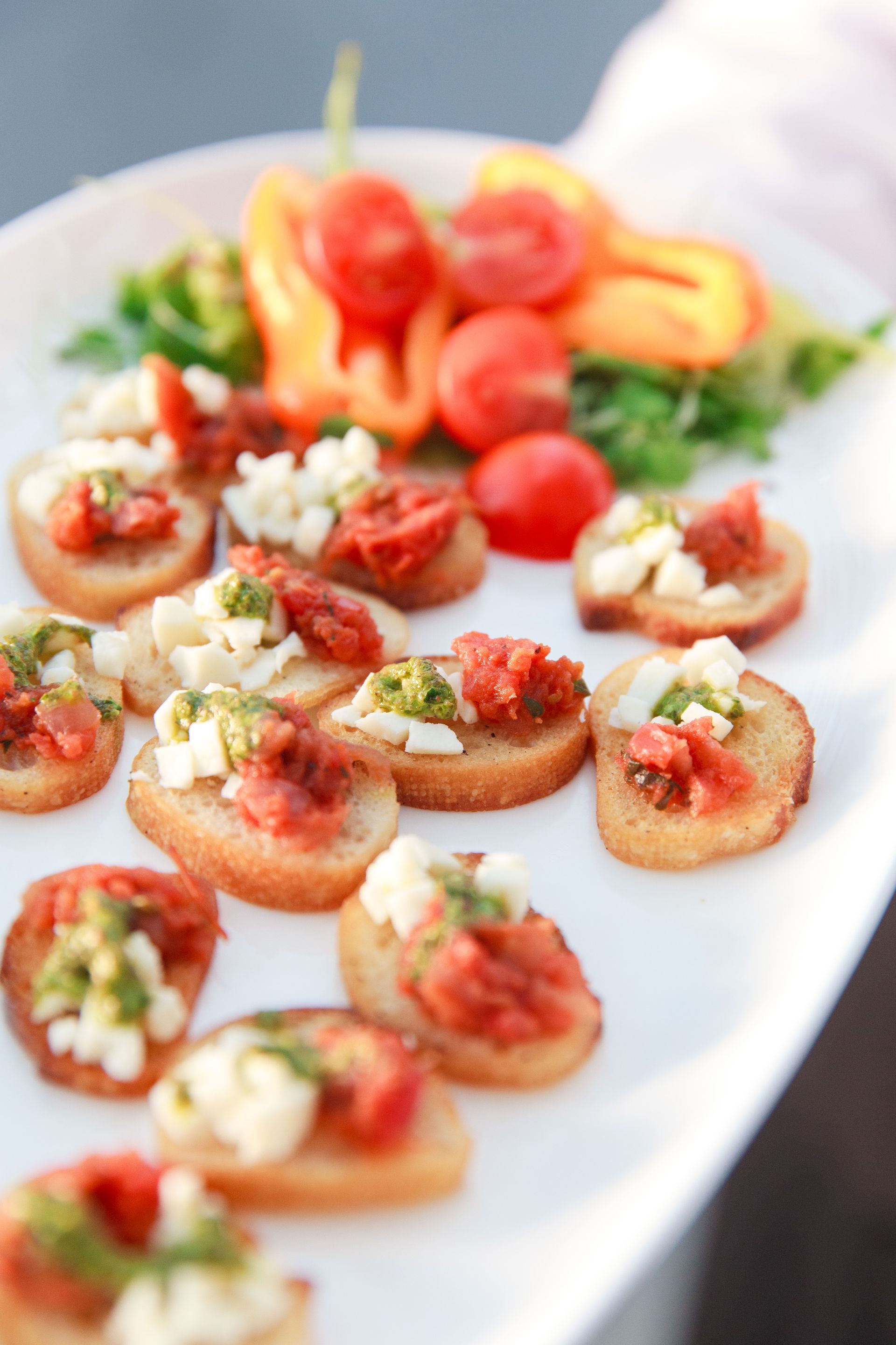 Bruschetta appetizers on a white platter, with tomatoes, peppers, and pesto.