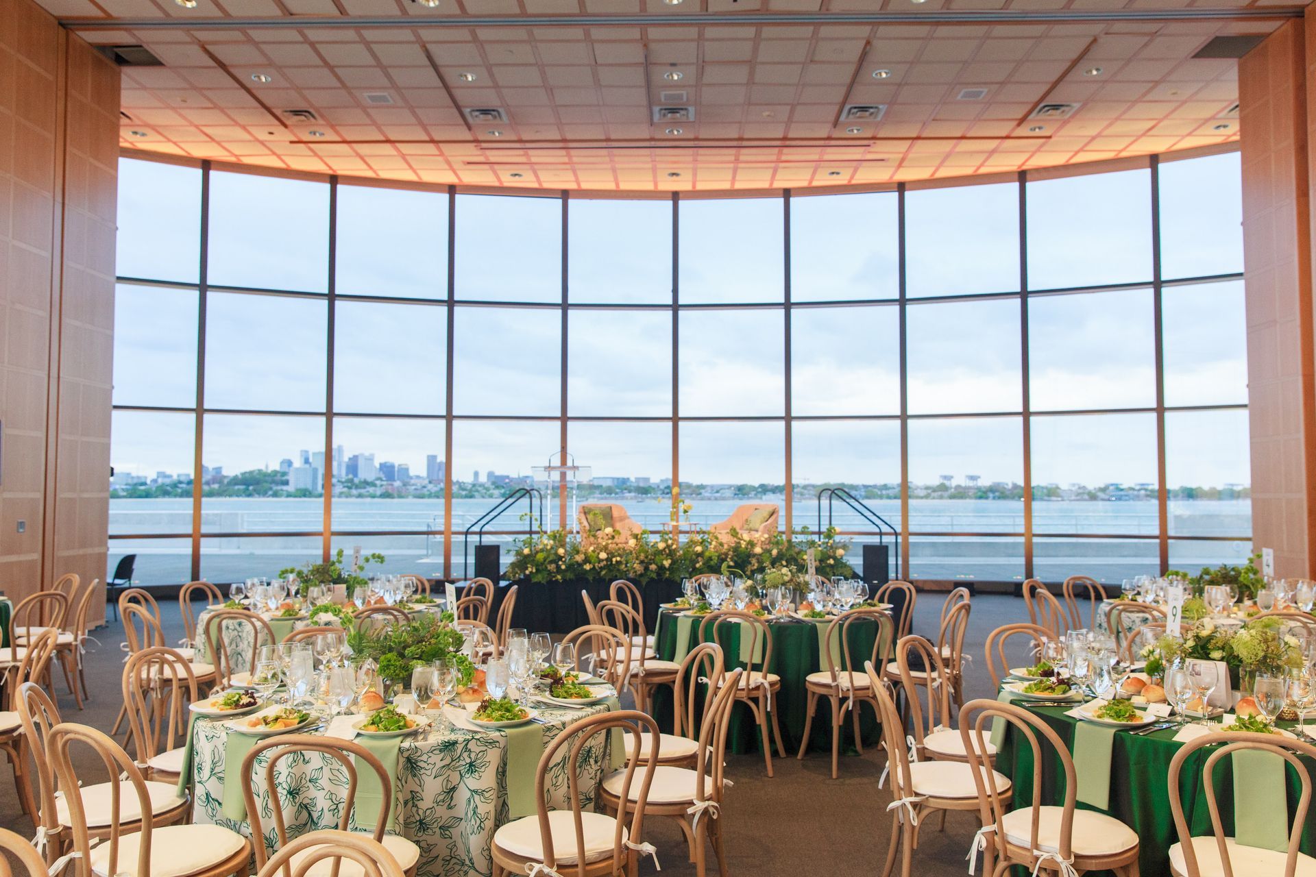 Elegant event space with round tables, green linens, wooden chairs, and a city view through curved windows.