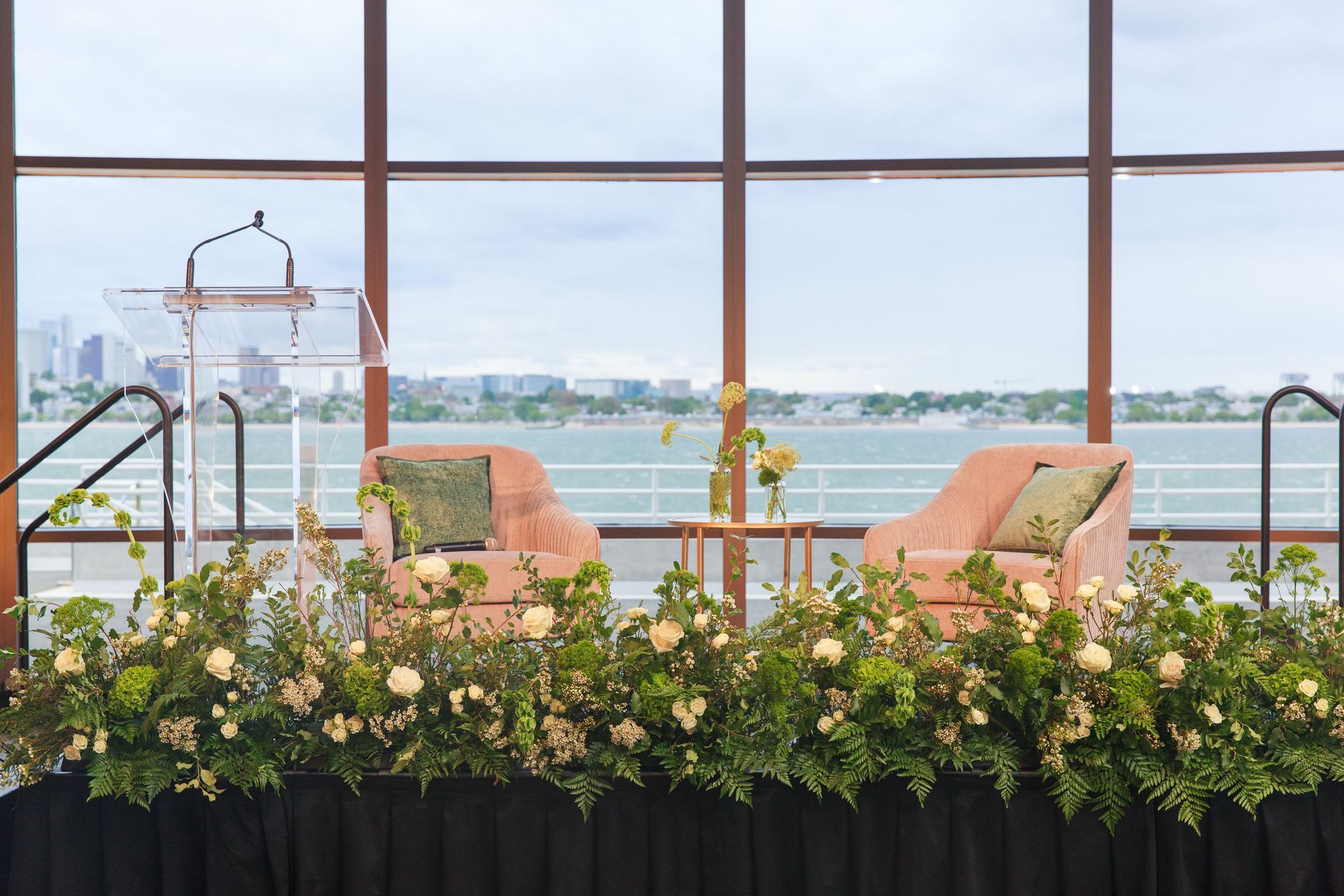 Podium, two pink chairs, and floral arrangements against a waterfront view.