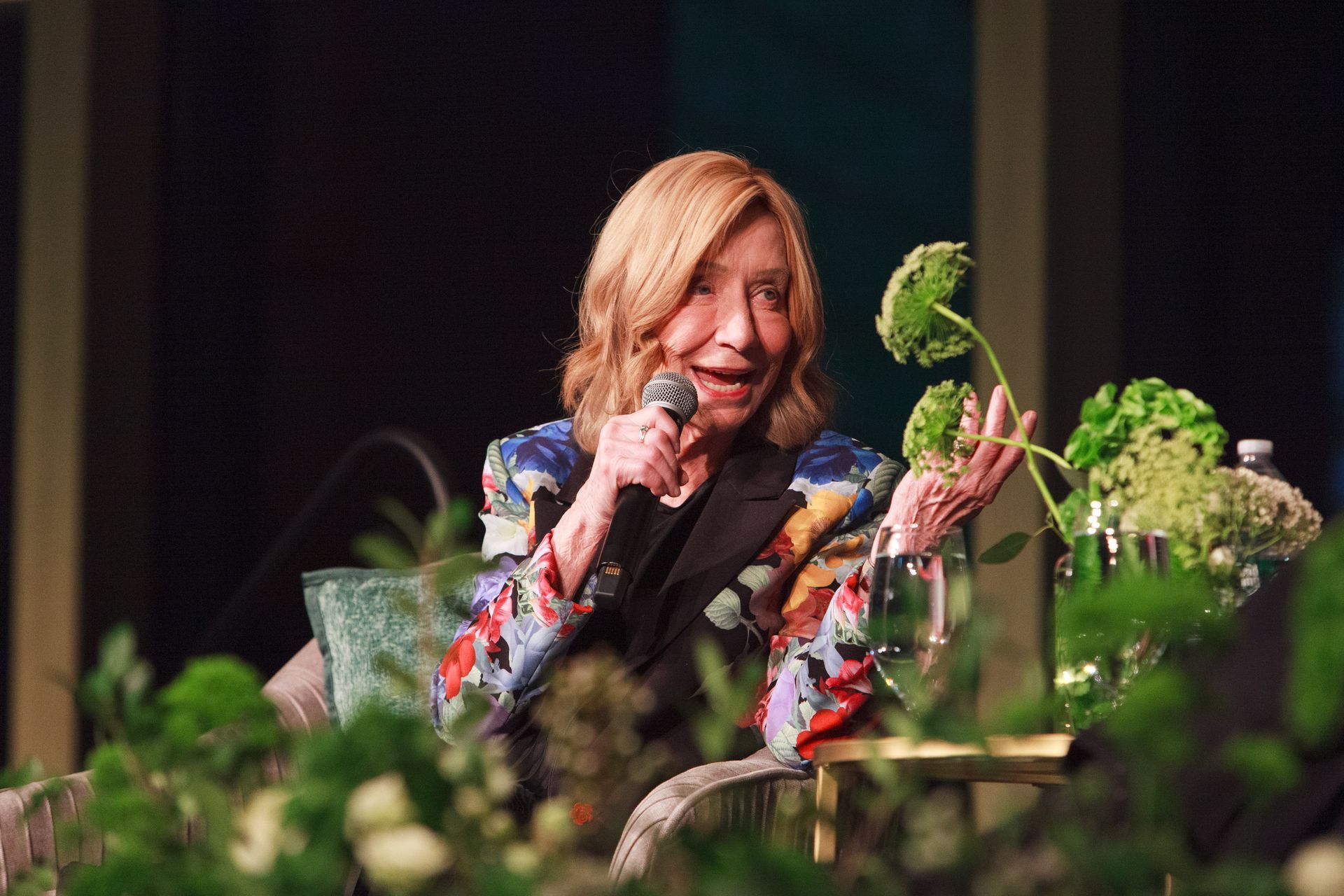 Woman with blonde hair speaks into a microphone, gesturing near greenery.