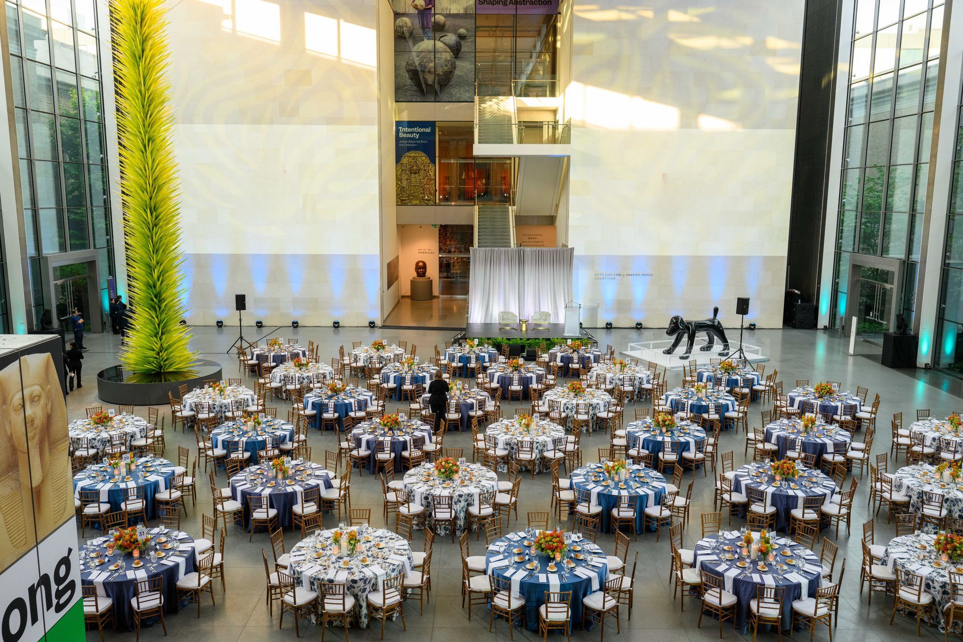 Formal event in a large, open hall with round tables set for a meal; a sculpture and tall flower arrangement are visible.