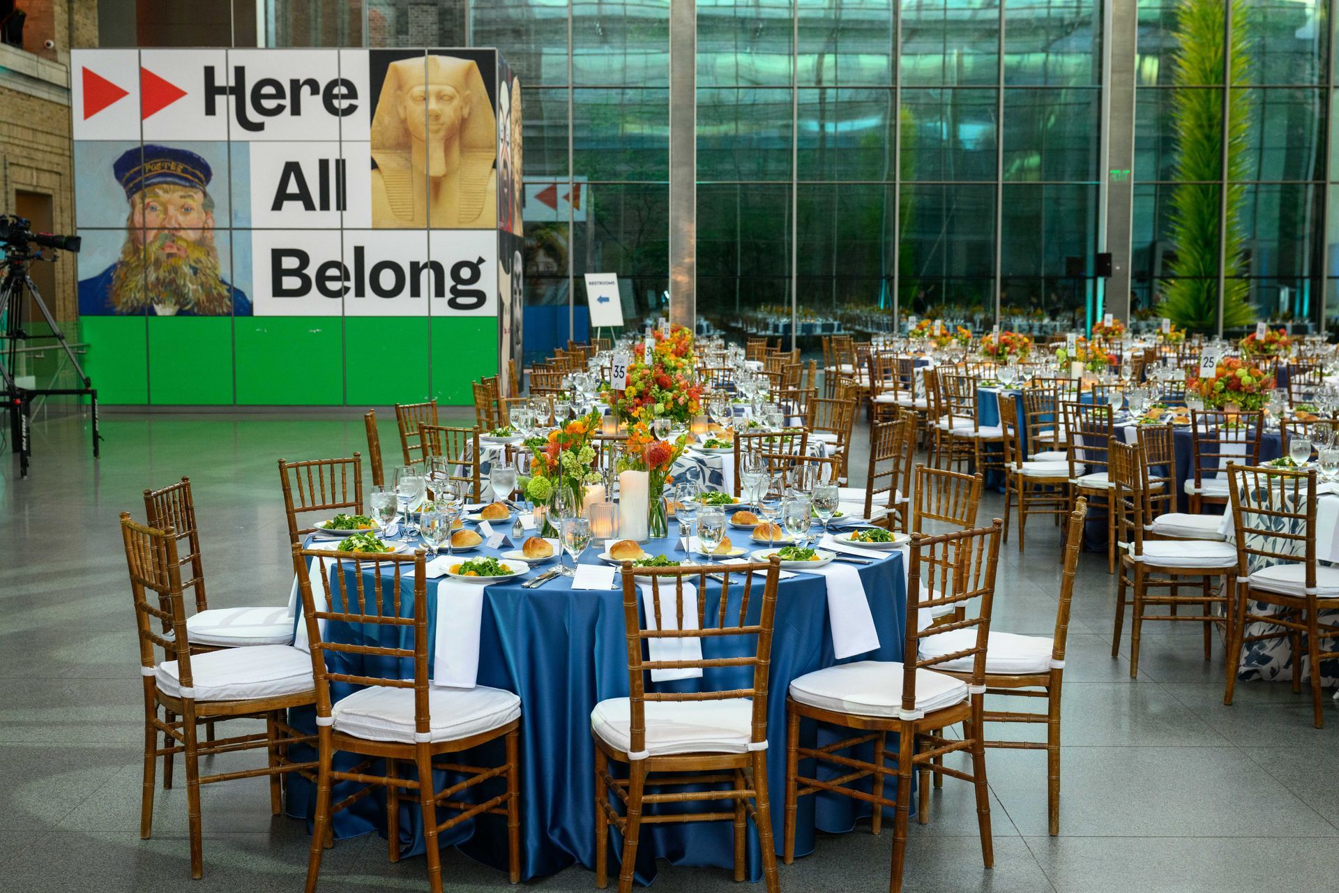 Round tables set for a formal event. Blue tablecloths, floral centerpieces, and wooden chairs. Large windows in background.