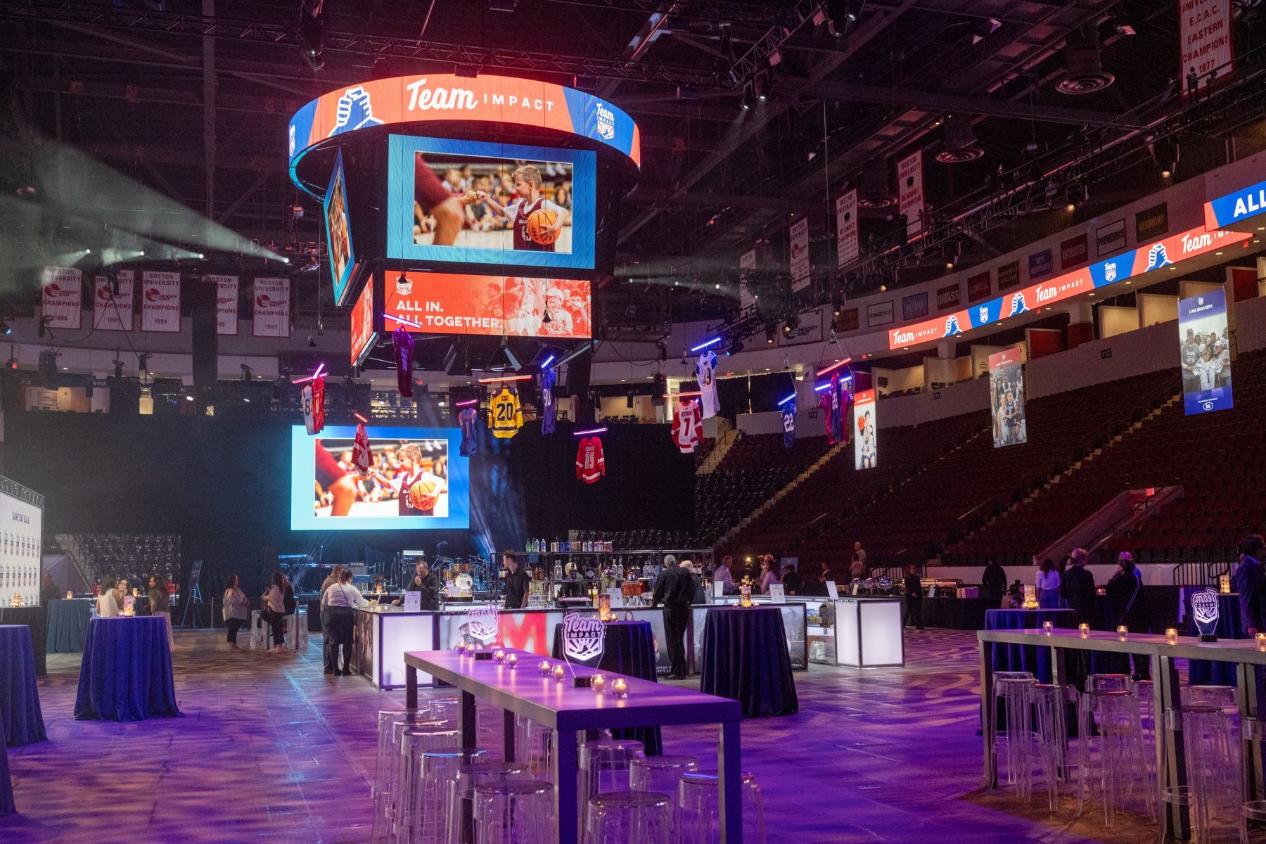 A large stadium with tables and chairs and a large screen hanging from the ceiling.