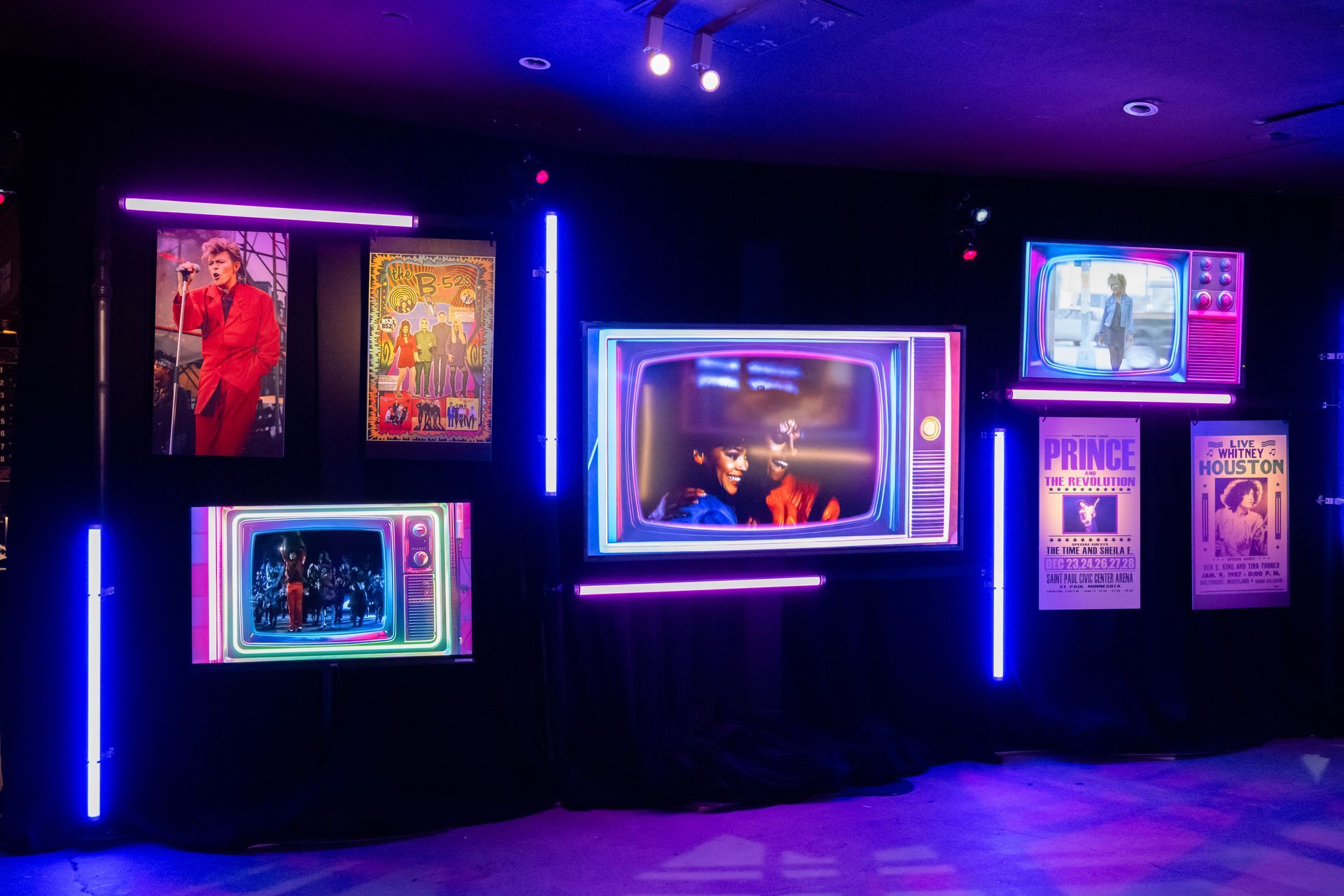 Wall display with purple and blue neon lights, framed TVs showing video content, posters.