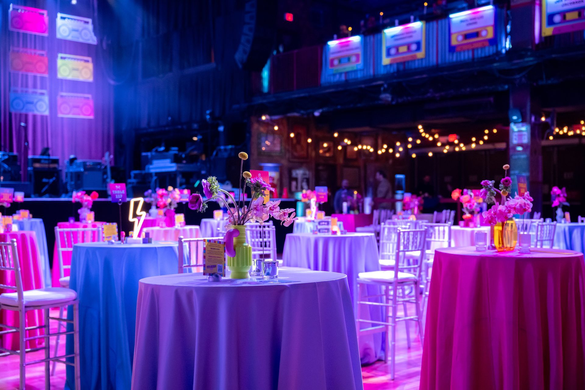 Tables with colorful linens in a brightly lit event space with a stage in the background.