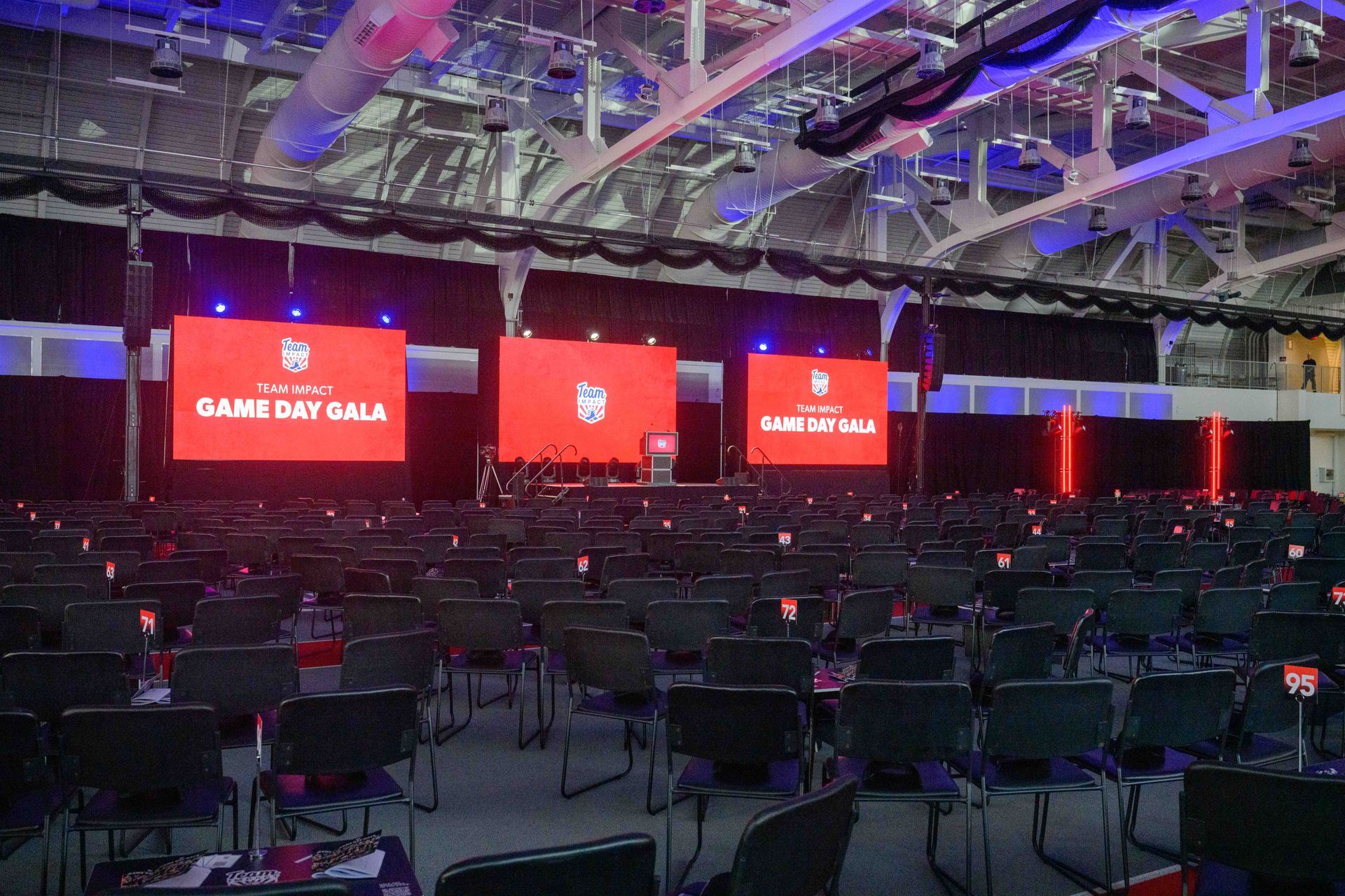 Empty auditorium set up for an event, with rows of chairs facing a stage with three screens.