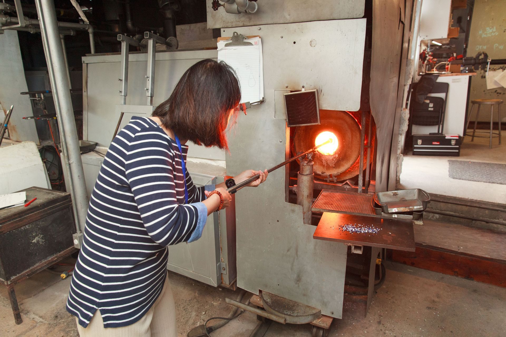 Woman heating glass in a furnace, using a rod. Workshop setting, bright orange glow, striped shirt.