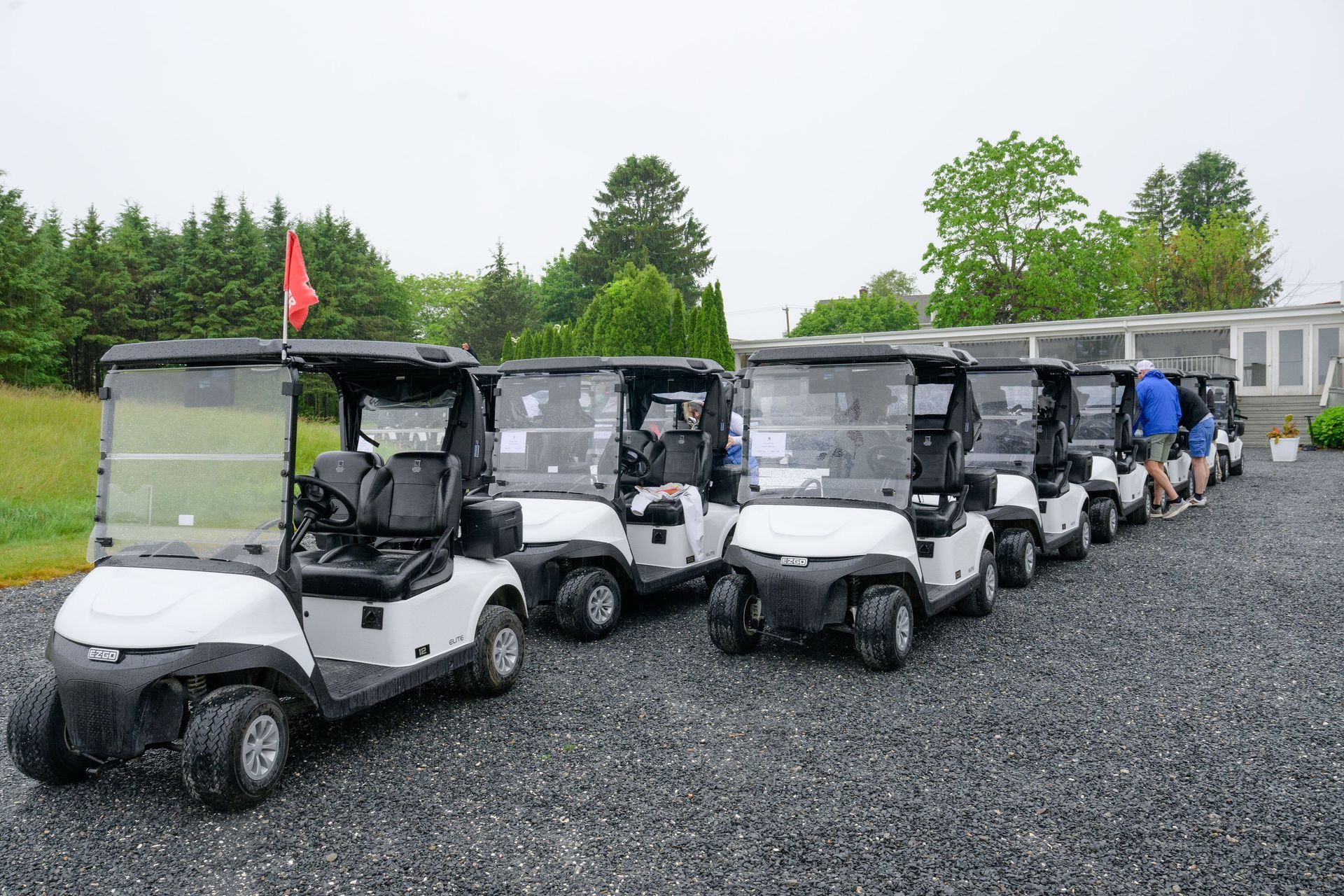 A row of white golf carts parked on gravel with people nearby, under overcast sky.
