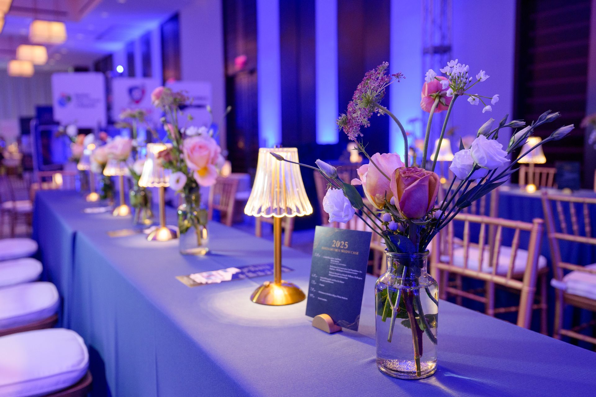 Long table set for event, blue tablecloth, flowers, small lamps. Soft blue lighting, chairs visible.
