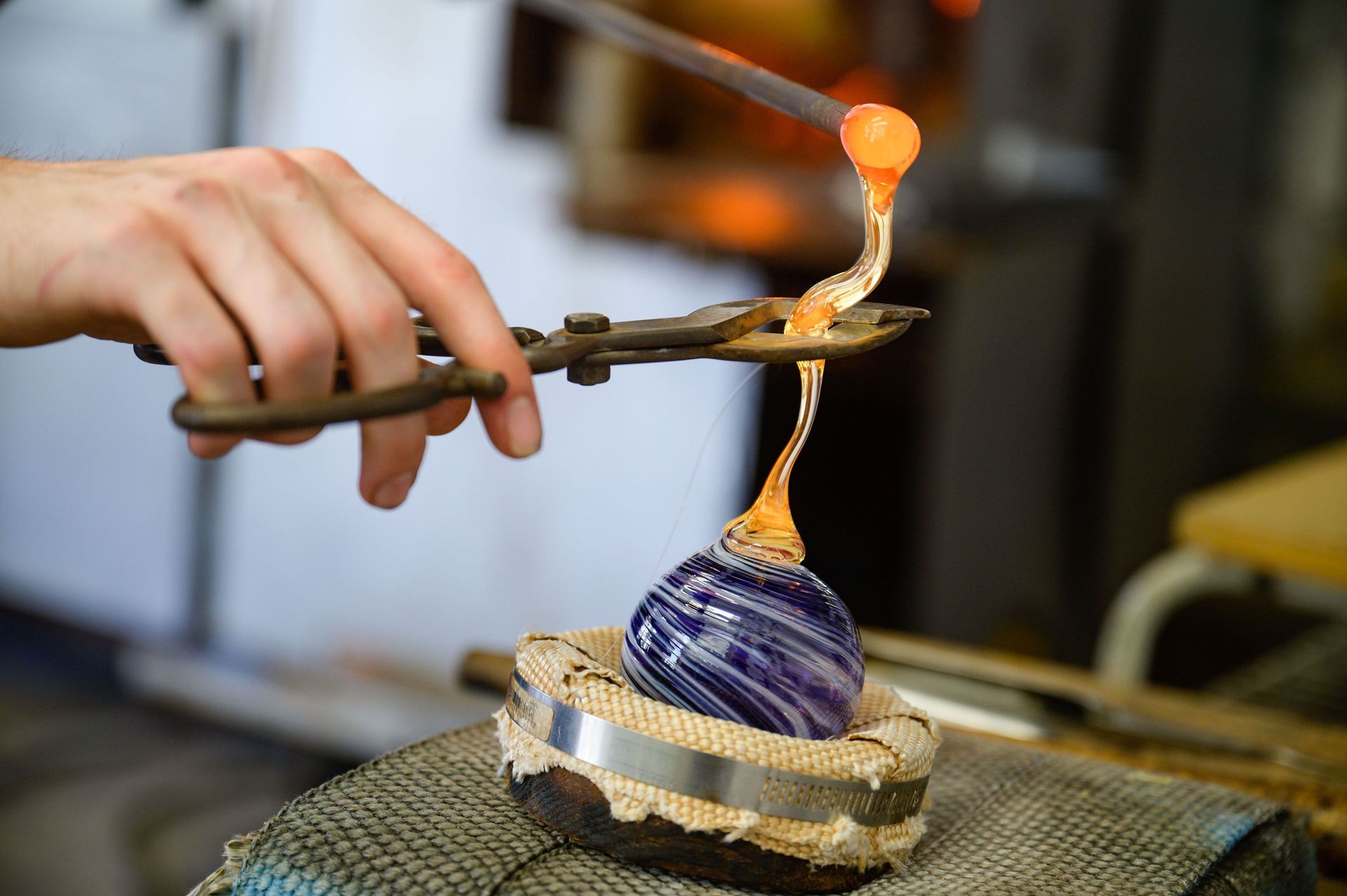 A person shaping molten glass with tools, adding a top to a purple and blue swirled glass object in a workshop.