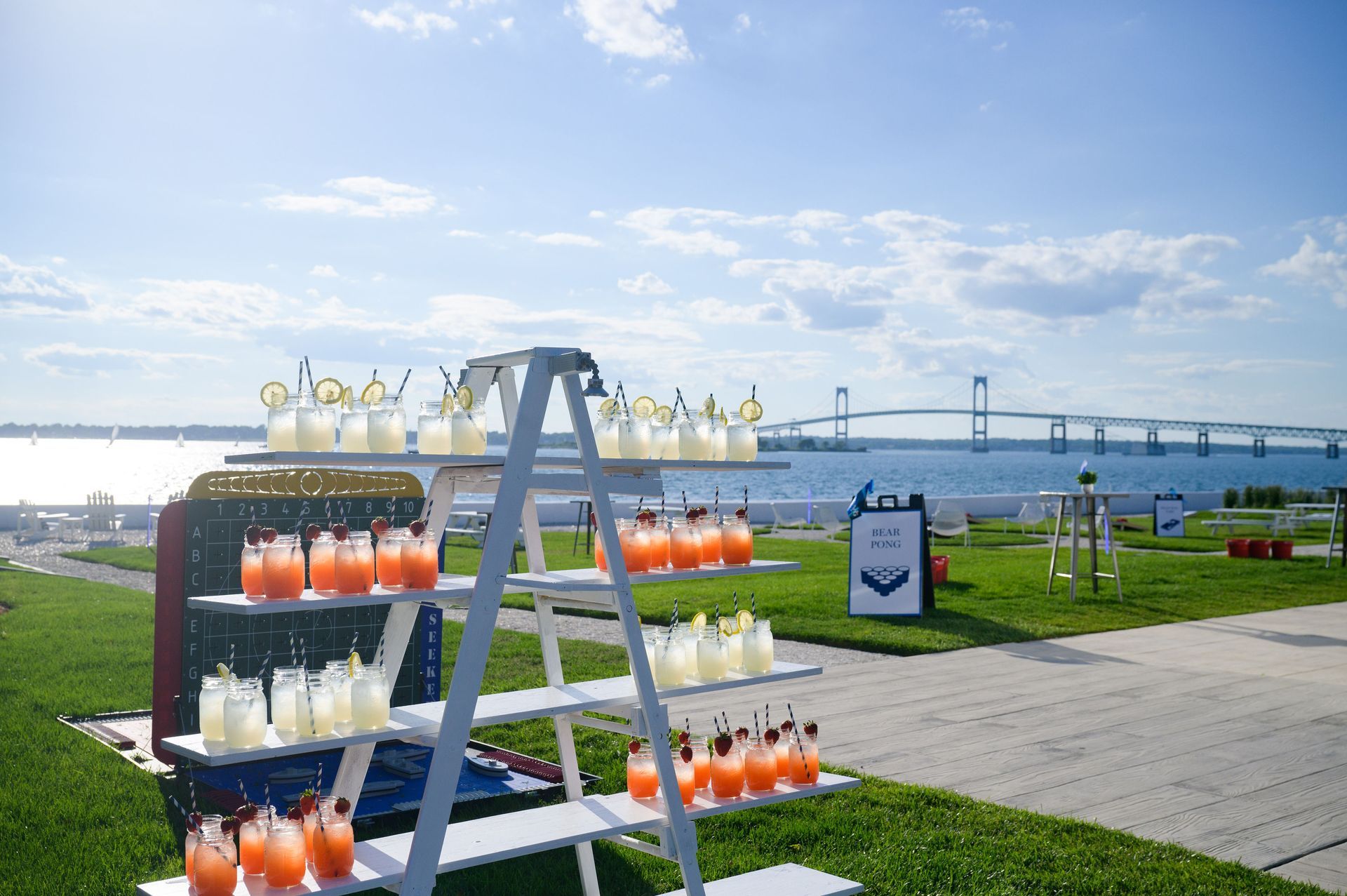Cocktail display with beach and bridge views: red & yellow drinks on white shelves, sunny day.