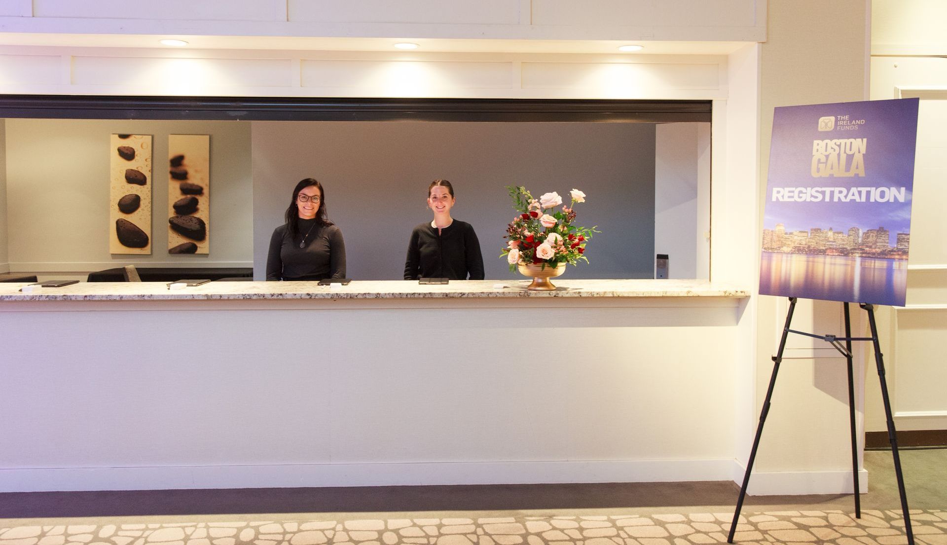 Two women are standing at a hotel reception desk.