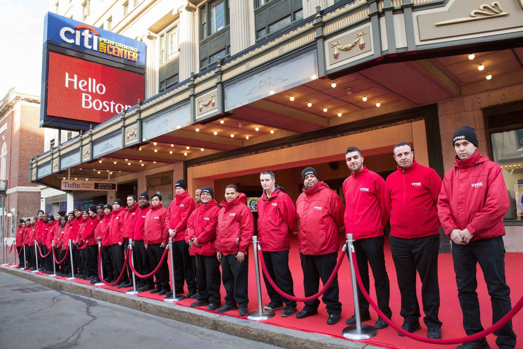 A group of men in red jackets are standing on a red carpet in front of a building.