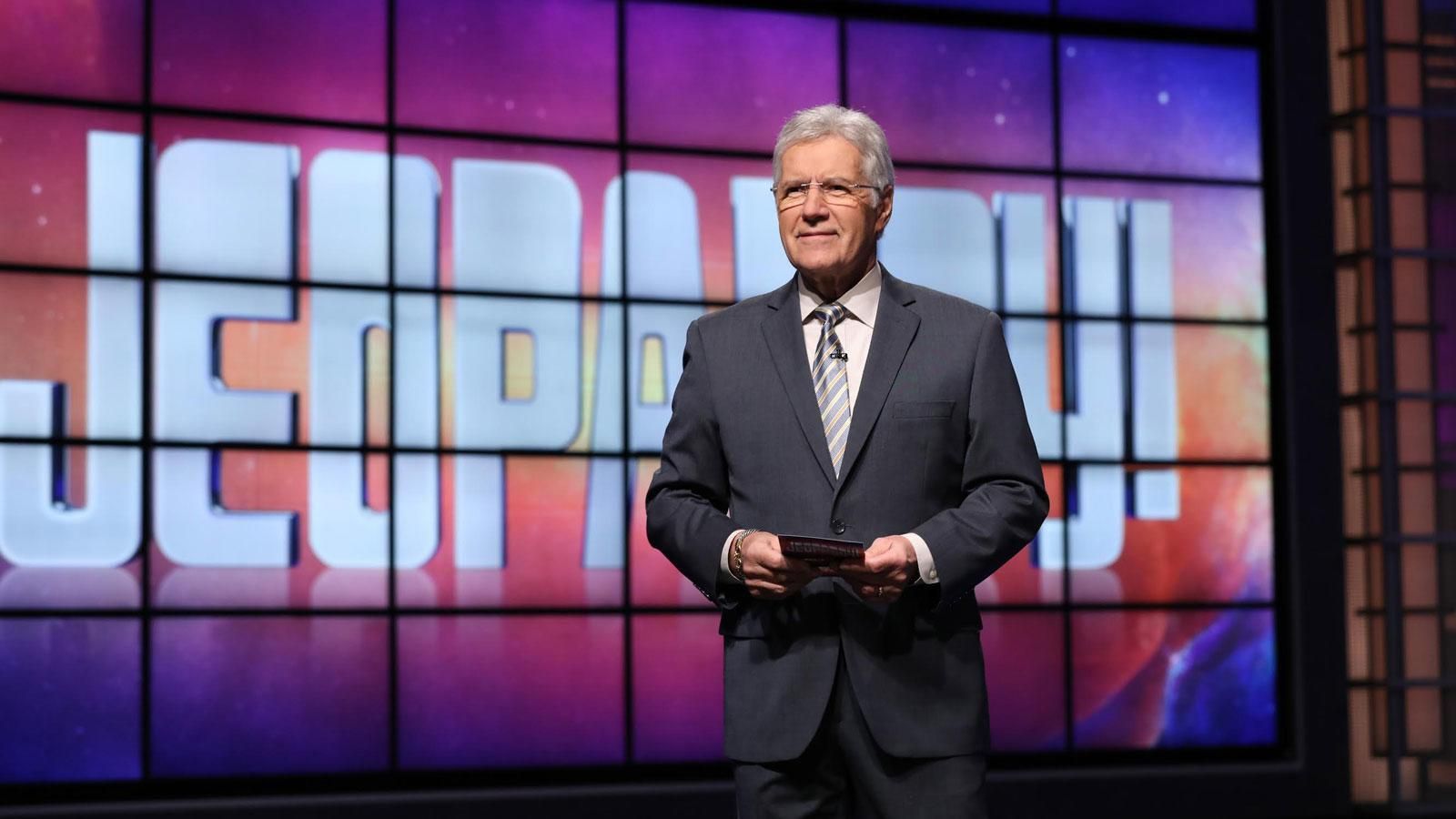 A man in a suit and tie is standing in front of a screen that says jeopardy