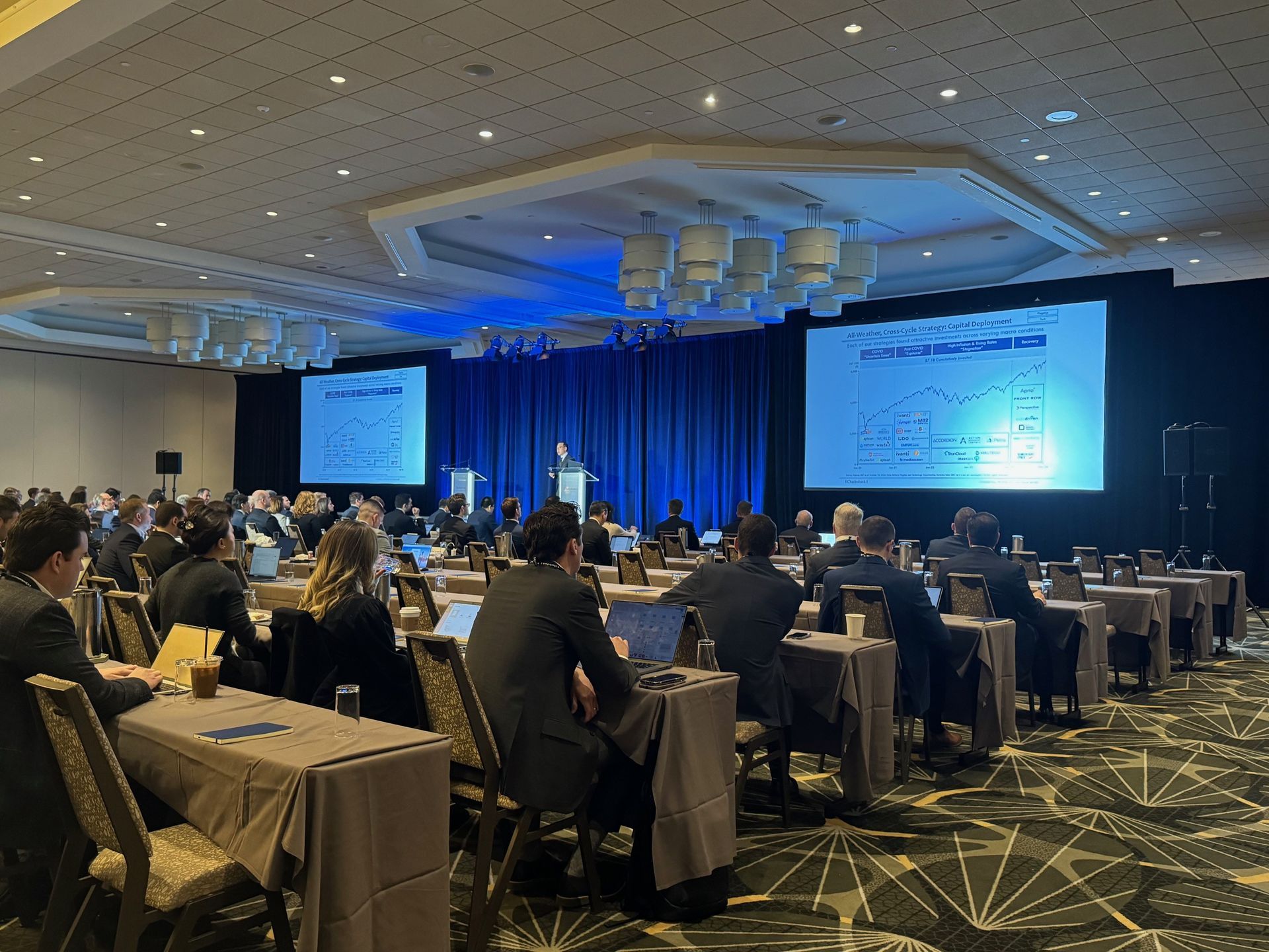 Conference in a large room, with attendees seated at tables, presenters on stage with screens.