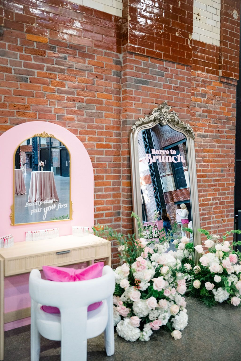 Pink vanity area with mirror, chair, and flowers against a brick wall.
