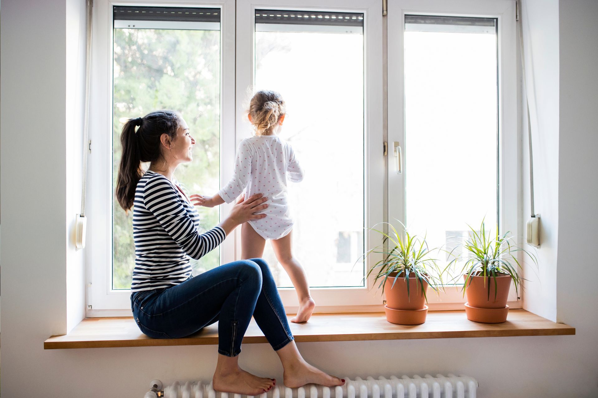 A woman and a little girl are sitting on a window sill looking out the window.