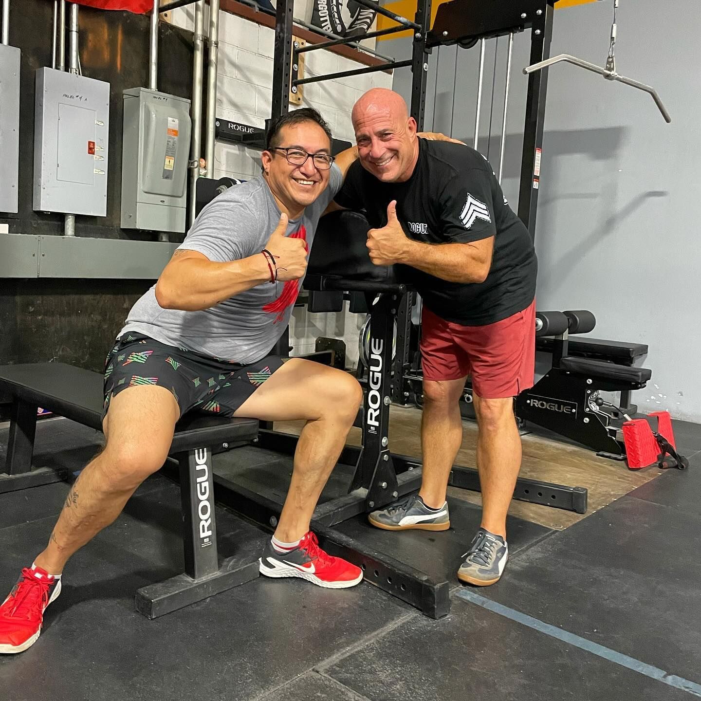 Two men are posing for a picture in a gym and one of them is giving a thumbs up.