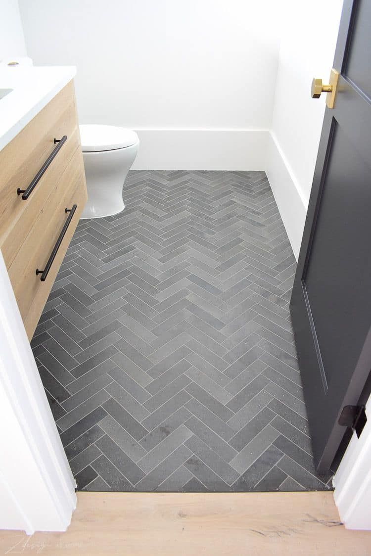 Narrow bathroom with gray herringbone tile floor, wood vanity, and black door.