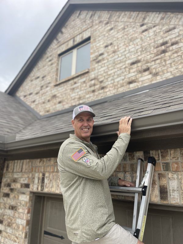 Man on a ladder, cleaning gutter on a brick house. He's smiling, wearing a hat and tan shirt.