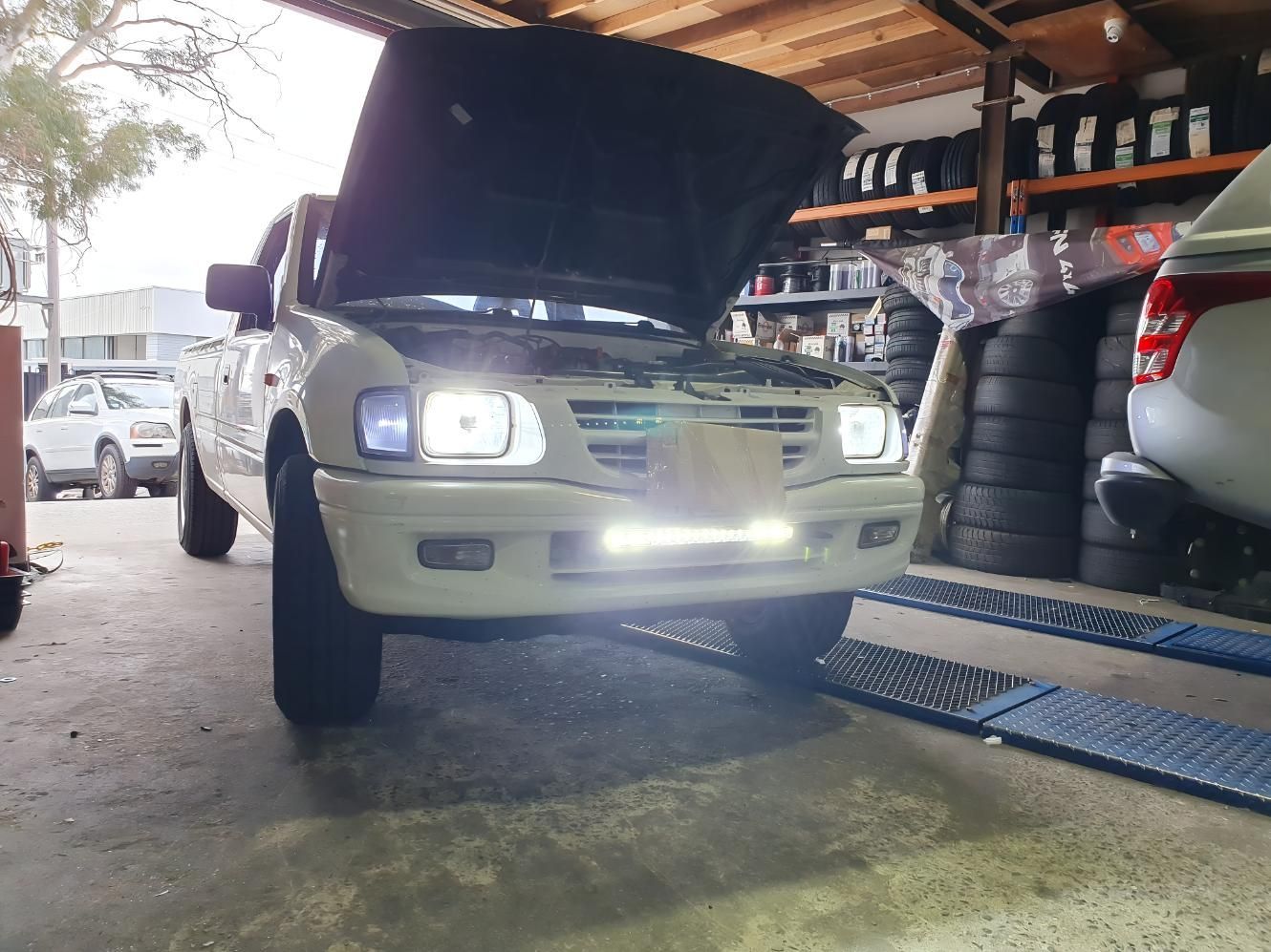 White Pickup Truck With Hood Open In A Garage, Illuminated By Headlights And A Light Bar