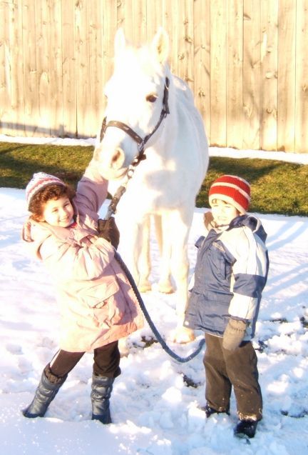 Two children in winter coats holding the lead rope of a white horse in snowy yard.