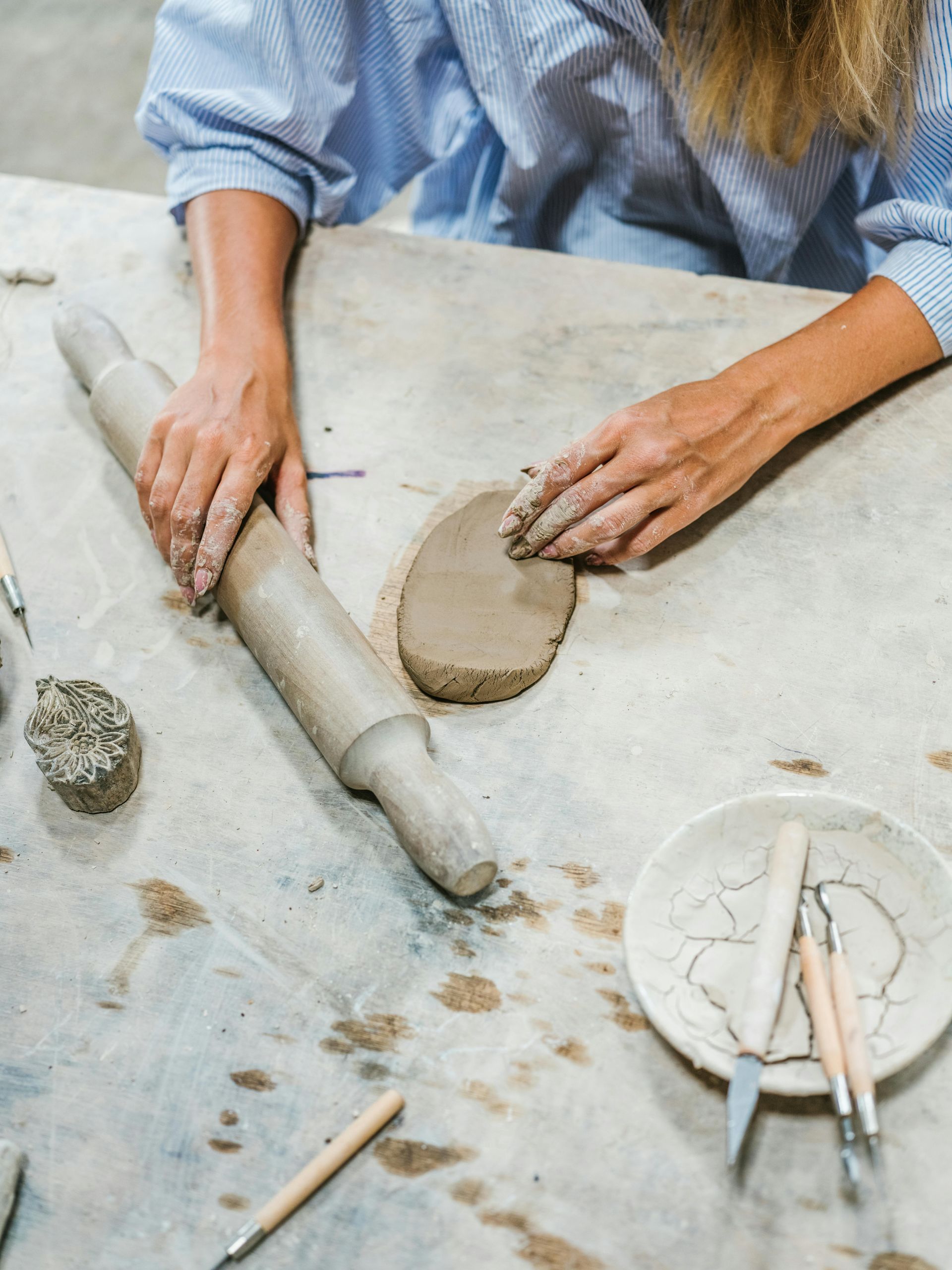 A person shapes a slab of clay with a rolling pin on a workshop table surrounded by pottery tools.