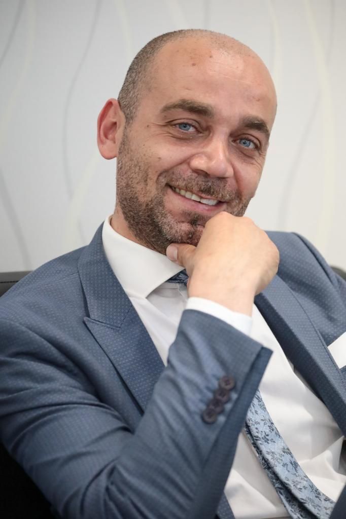 Uomo in abito blu, mano sul mento, sorridente. Camicia bianca, cravatta fantasia, fazzoletto da taschino. Ambiente interno.