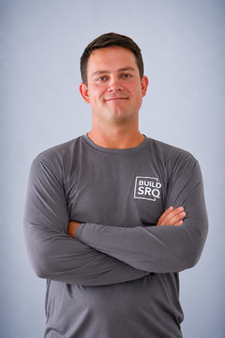 Man in a gray shirt with arms crossed, standing against a light blue background.
