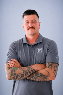 Man with mustache and tattooed arms wearing a gray polo, standing with arms crossed against a plain blue background