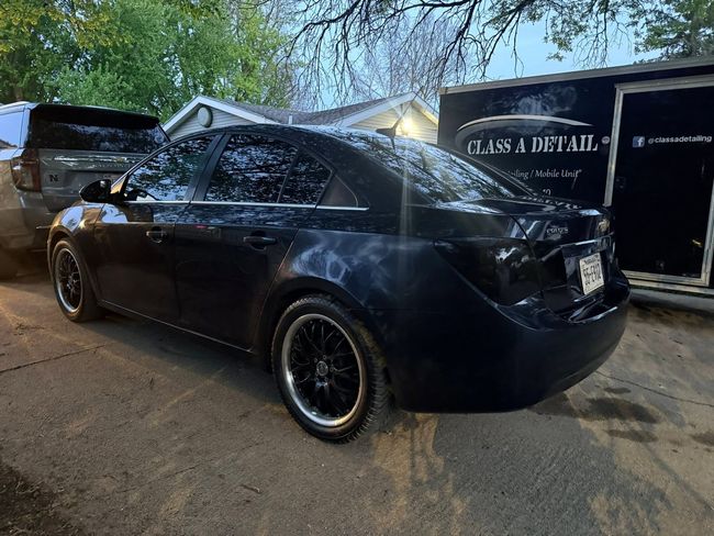 Black sedan with rear-end damage parked beside a trailer, with water spraying over it at dusk.