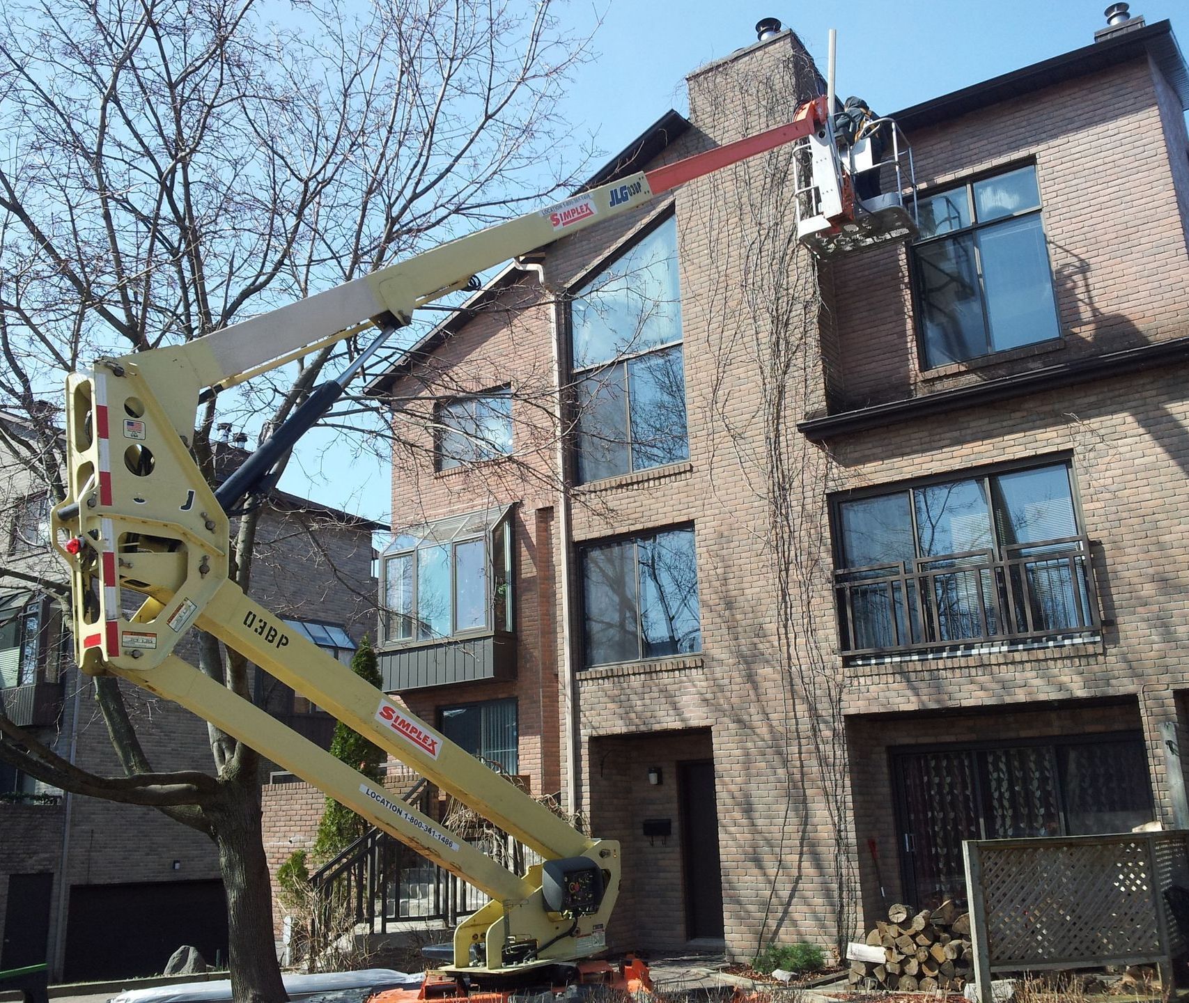 Une nacelle élévatrice jaune s'étend vers un bâtiment en briques. Des travaux de cheminée sont en cours.
