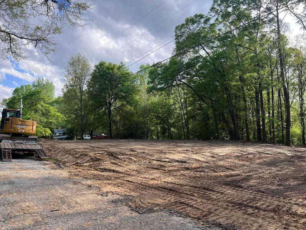 A construction site with a large cleared dirt area, excavator, and trees in the background.