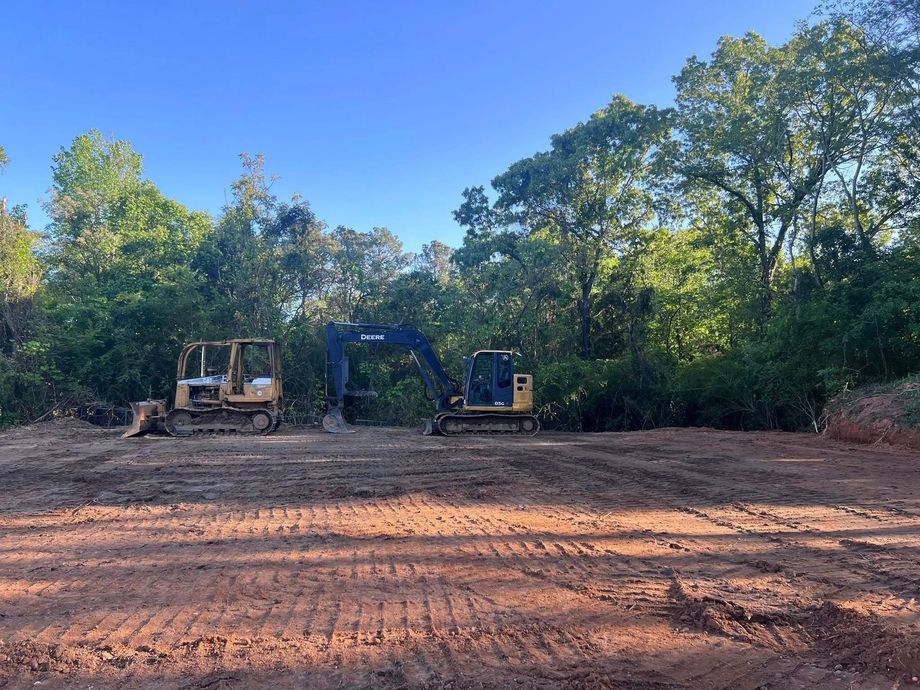 Construction site: Bulldozer and excavator on cleared land, trees in the background, blue sky.