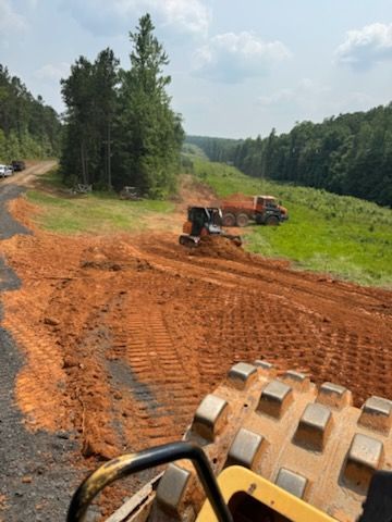 Construction site with a roller compactor in the foreground, earthmoving equipment, and a truck. Red dirt and green vegetation.
