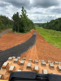 Road construction, red dirt and dark asphalt path through a grassy area, seen from a construction vehicle.