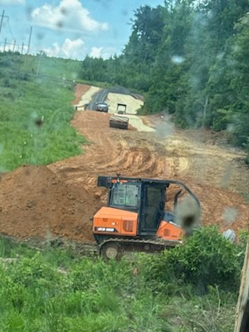 Orange bulldozer on dirt road construction site; two rollers and a car visible up the hill, green forest backdrop.