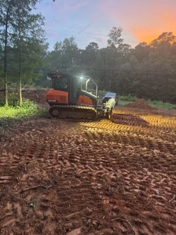 An orange track loader works on a muddy construction site at dusk with a sunset in the background.