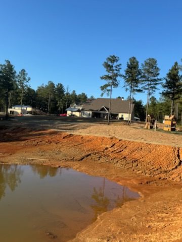 Construction site with pond in foreground, house in background, trees, and blue sky.