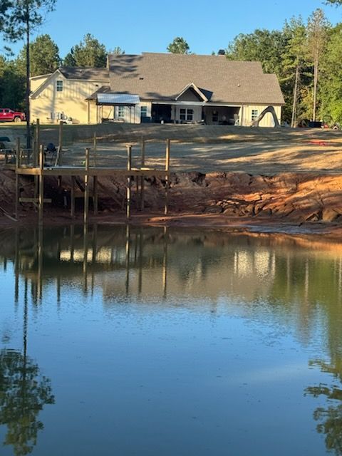 A house on a bank overlooks a pond with a wooden dock. The sky and trees reflect on the water.