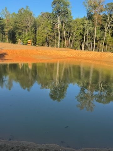 Reflective pond with tree reflections, orange bank, and treeline under a blue sky.