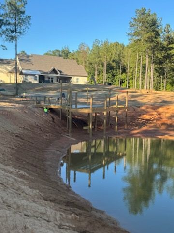 A wooden dock extends over a pond, reflecting its structure. A house and trees are in the background.