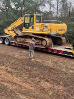 Yellow excavator on a trailer, man in camouflage stands beside it in a field.