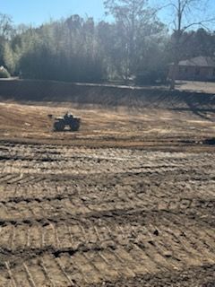 An ATV driving on graded, brown dirt with trees and a building in the background.
