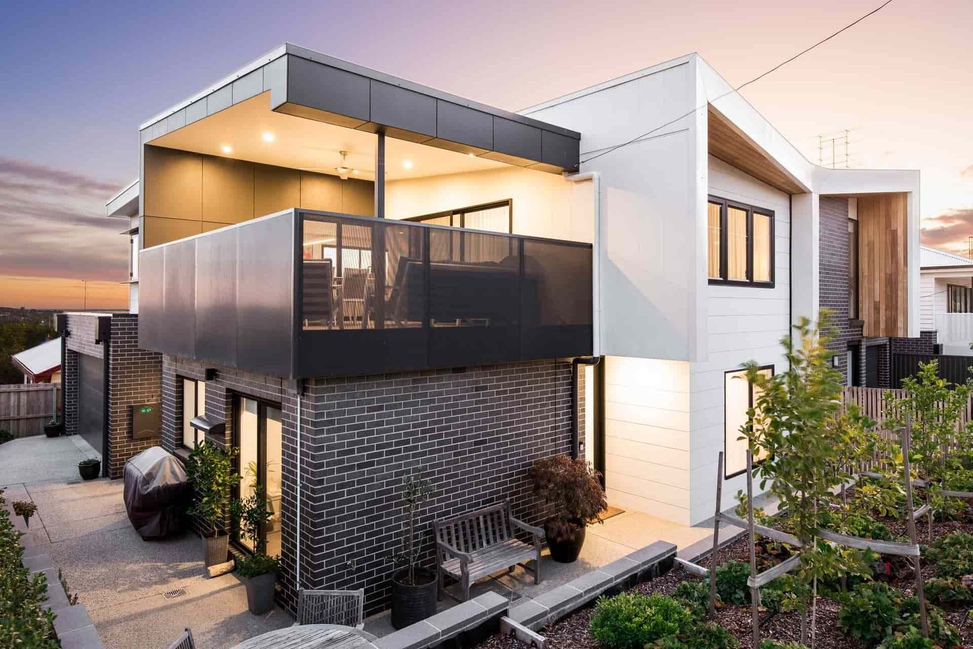 Modern two-story house with balcony and dark brick facade, with plants and sunset sky.