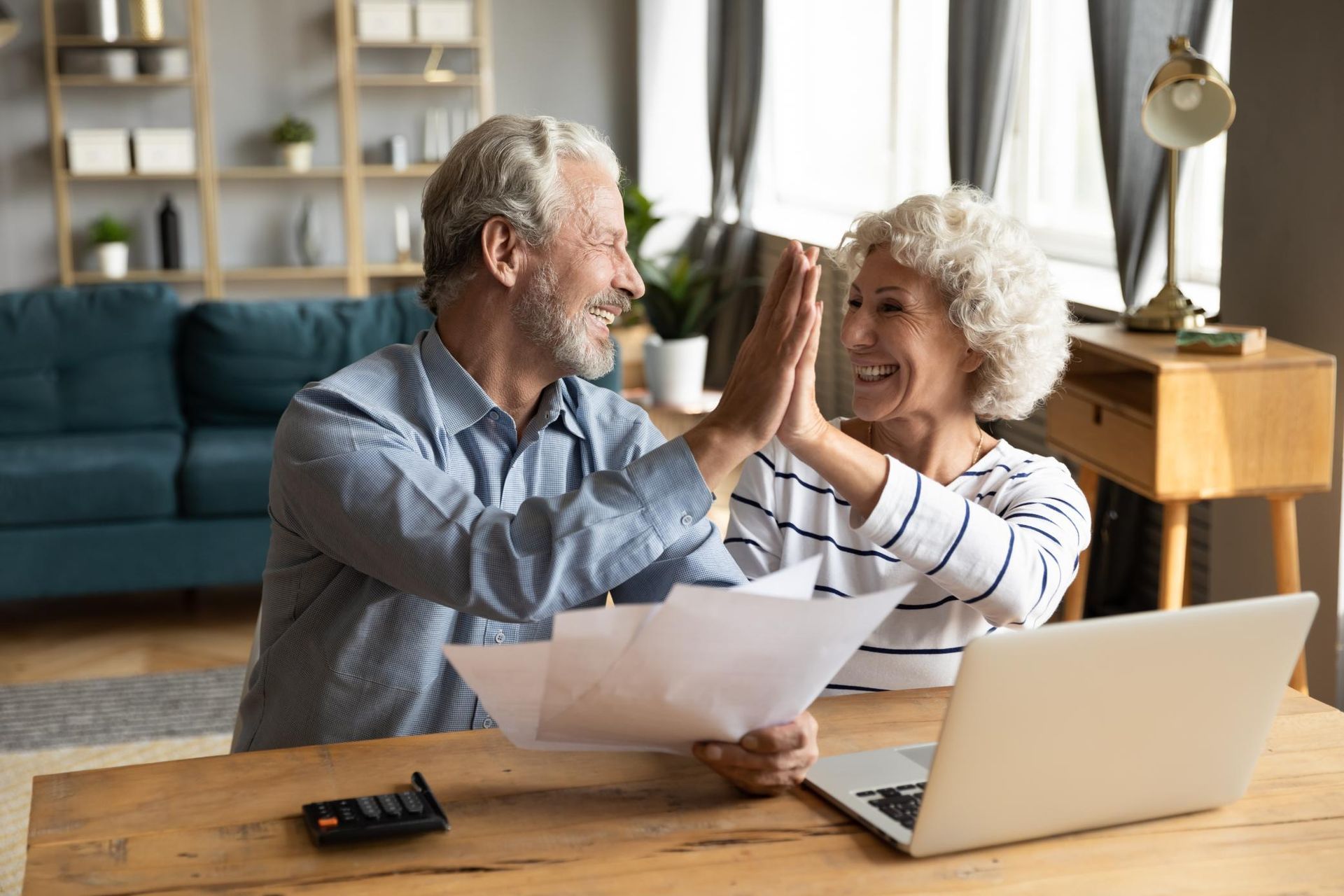 Senior couple happily high-fiving over paperwork and laptop at a table in their home.